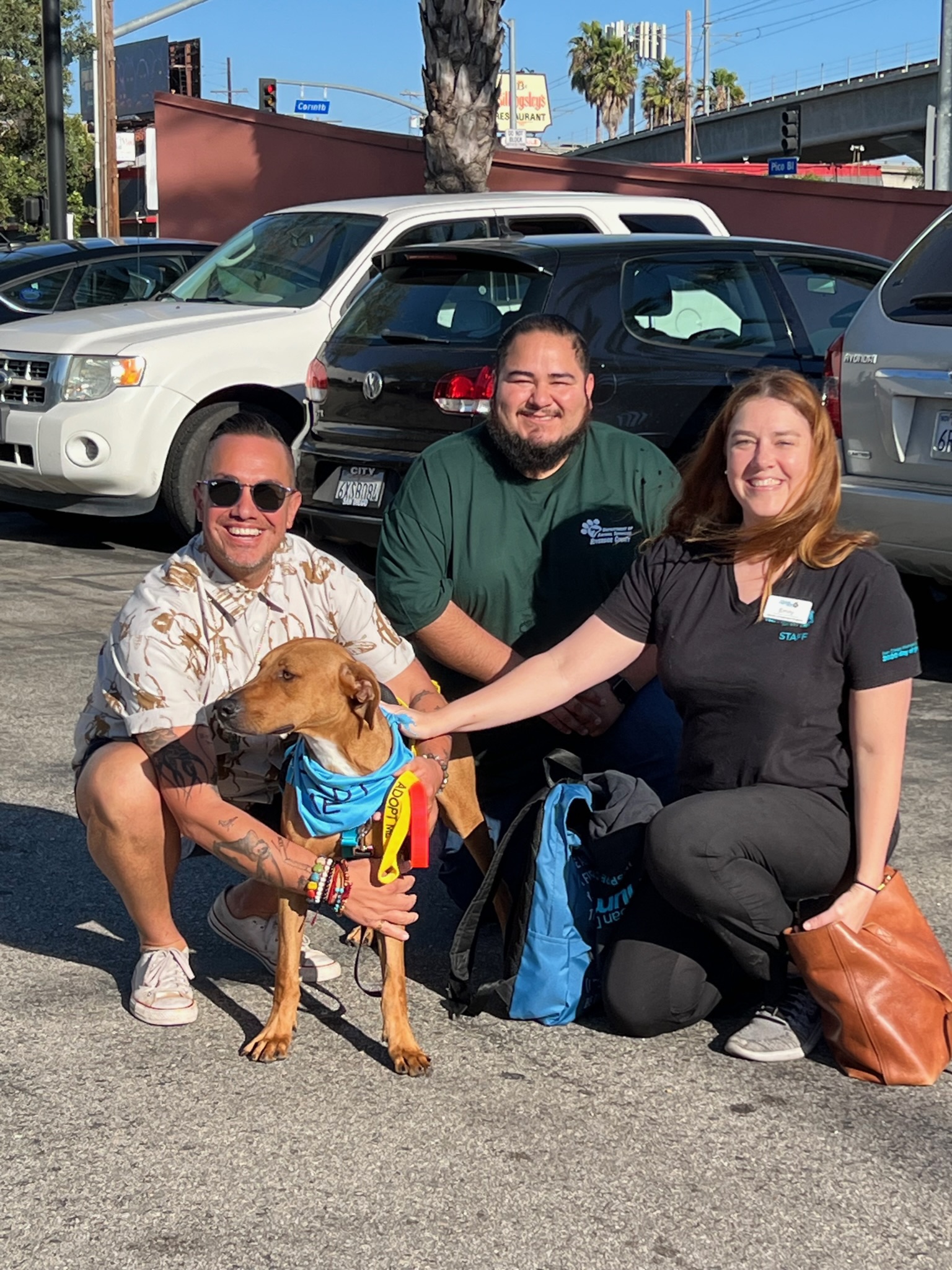 three people surround a brown dog wearing a blue bandana